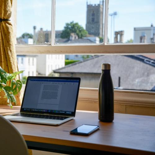 En dator och en vattenflaska placerade på ett skrivbord framför ett fönster.