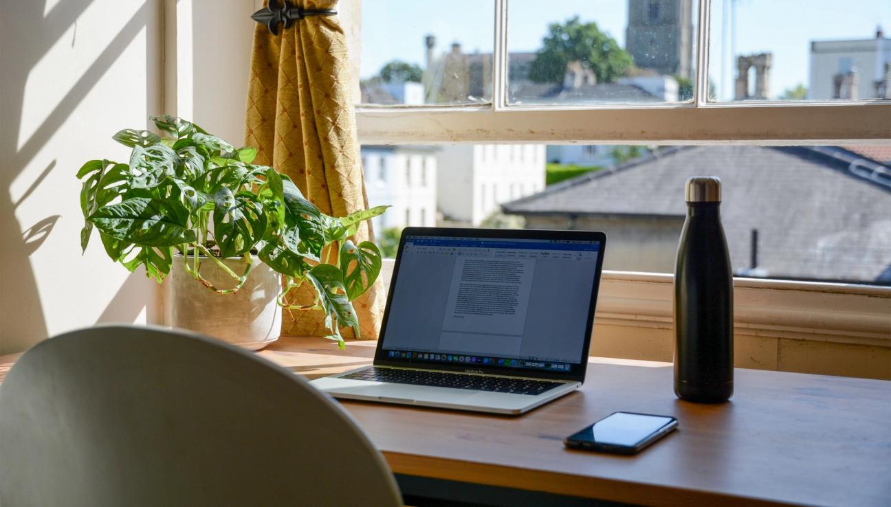 Pc, mobil, en plante og vannflaske på en pult foran vindu.