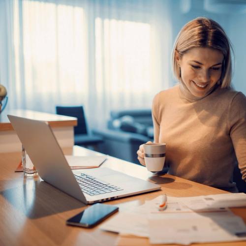 En kvinna sitter och ser över sina fakturor. Hon har pc och mobil på bordet framför sig. bild.