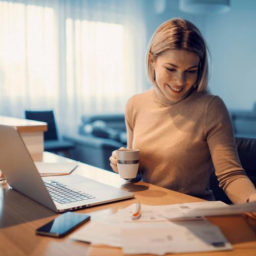 En kvinna sitter och ser över sina fakturor. Hon har pc och mobil på bordet framför sig. bild.