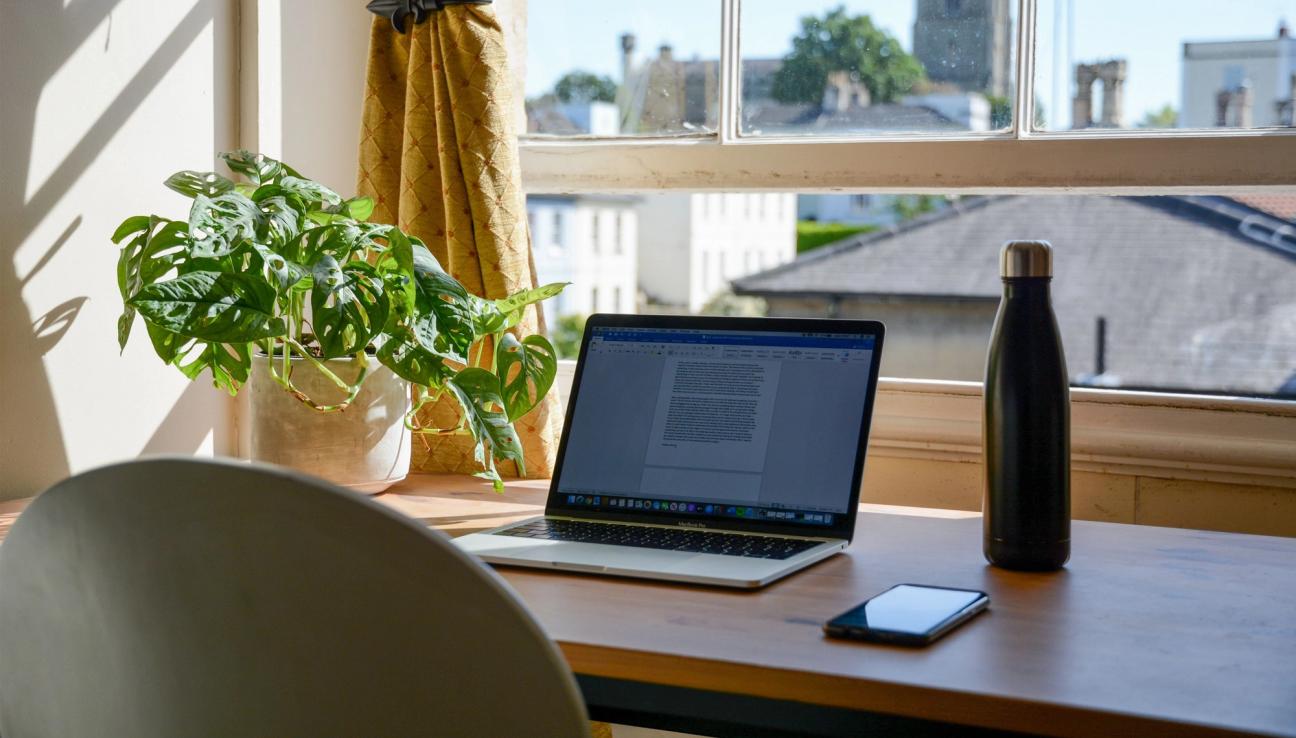 Tietokone, matkapuhelin ja vesipullo pöydällä ikkunan edessä. Valokuva.