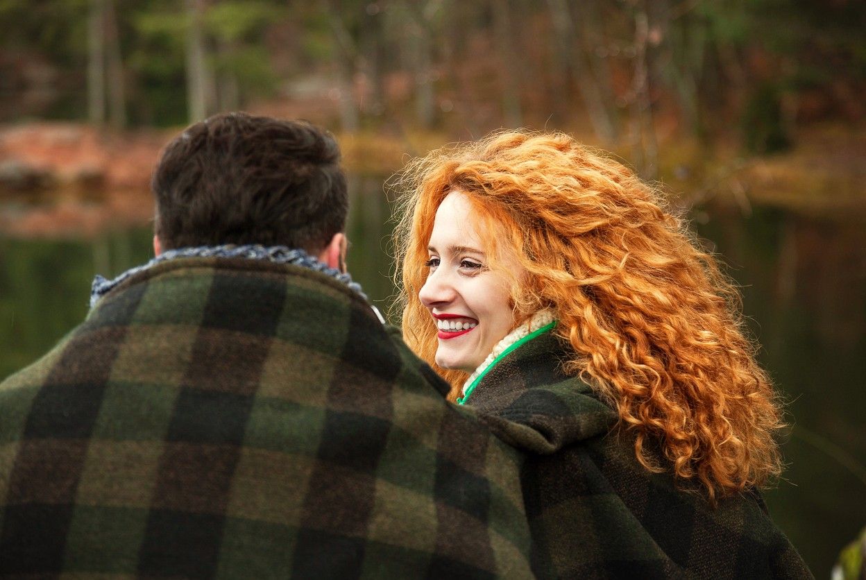 Mies ja nainen istuvat ulkona, viltti ympärillään. Naisella on kiharat punaiset hiukset ja leveä hymy kasvoillaan. Valokuva.