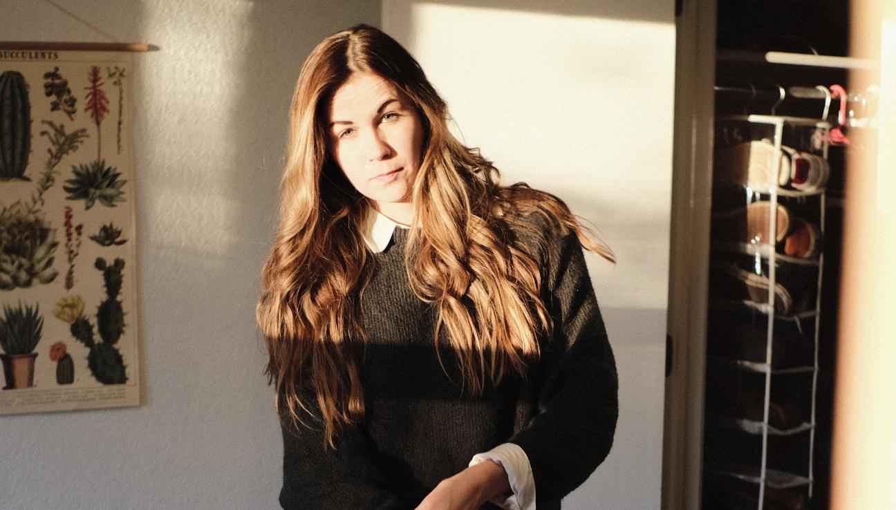 En kvinna poserar för kameran i solljuset. Hon håller på att vika upp ärmarna på sin skjorta.