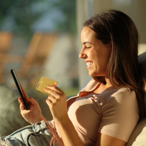 Dame sitter i sollyys med Morrow kredittkort i ene hånden og mobilen i den andre. bilde.