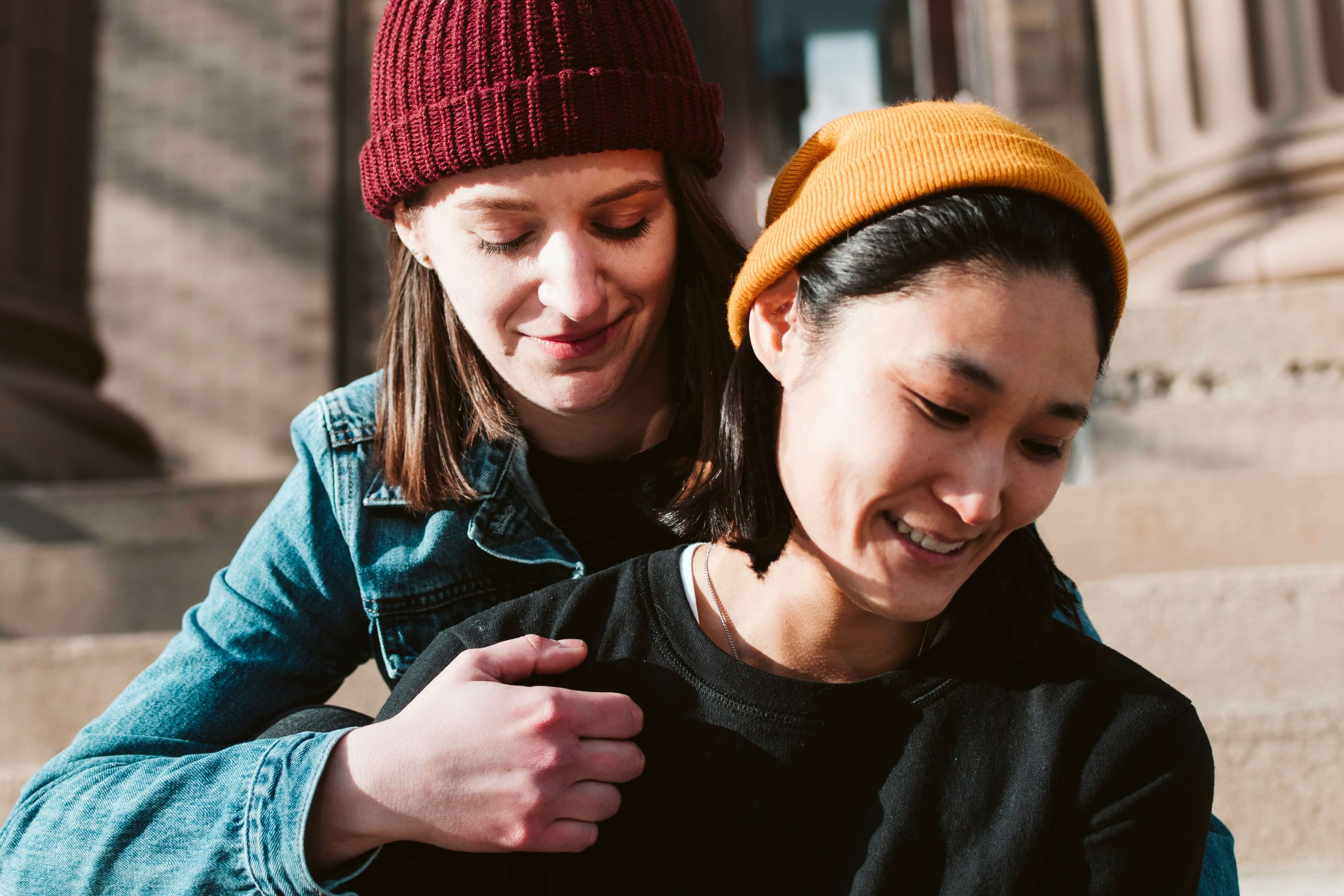 To kvinner sitter tett sammen. Den ene har rød lue og den andre har gul. Høstsolen skinner. bilde.