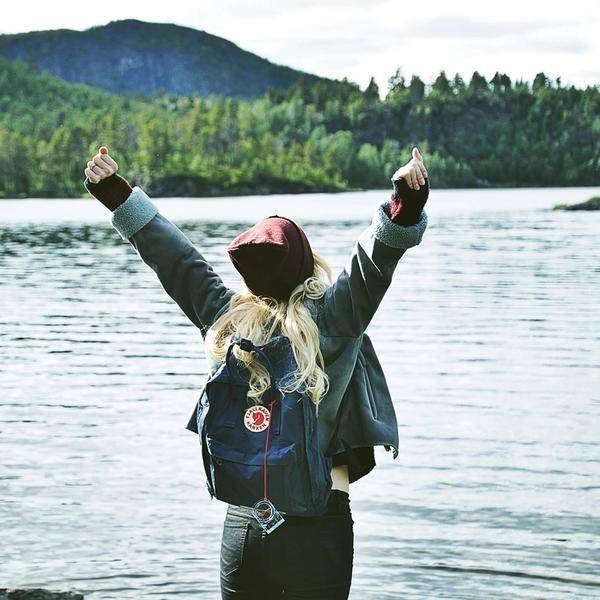 En kvinna med Fjällrävensäck står vid en skogssjö och sträcker armarna i vädret.
