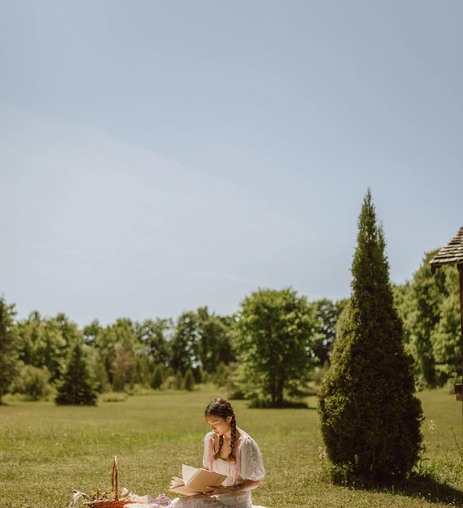 A girl with braids and a pretty dress, reading in a big grass field.