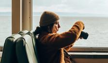 A girl sitting on a train taking pictures of the lake outside the window.