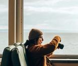 A girl sitting on a train taking pictures of the lake outside the window.