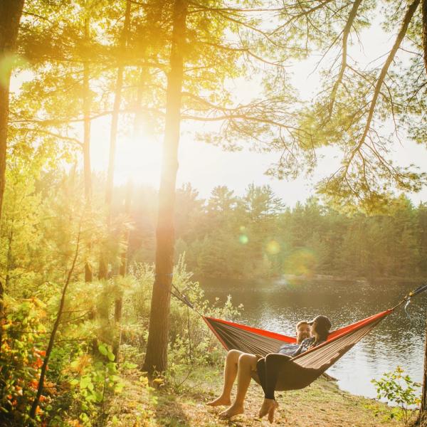A couple is sitting in a hammock by a lake. The sun is shining.