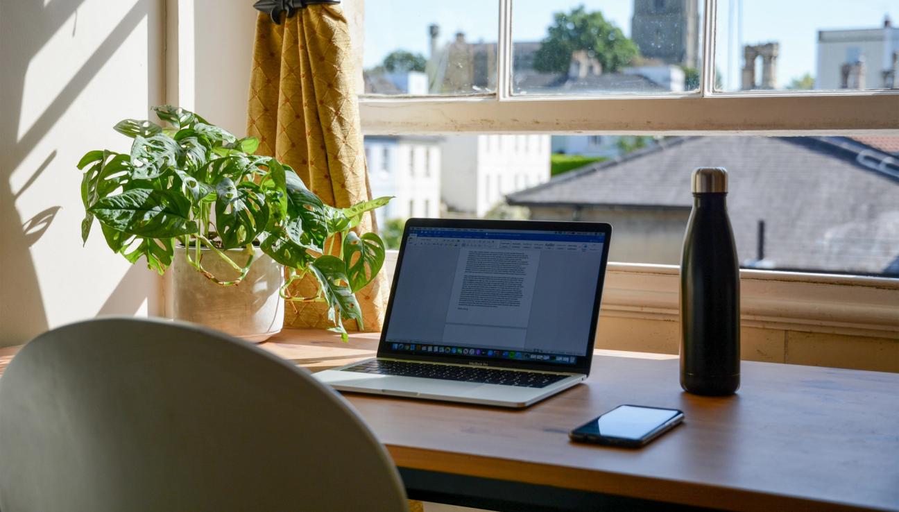 A computer and a waterbottle placed on a desk in front of a window.