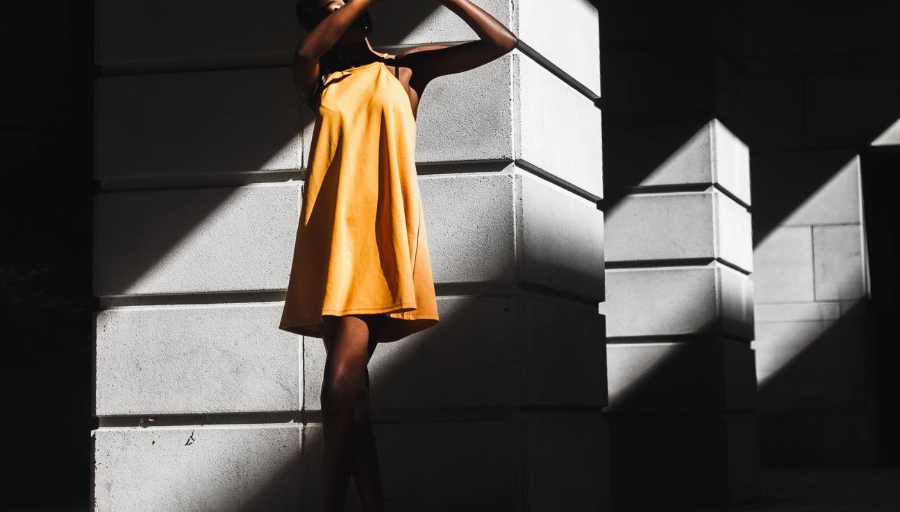 Woman in a yellow dress standing in direct sunlight and covering her eyes from the sun.