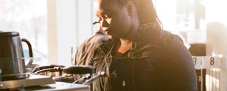 A barista that is working behind the counter. The sun is shining at the back of her head.