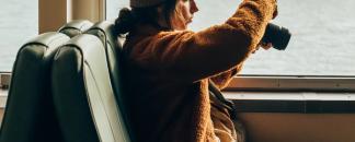 A girl sitting on a train taking pictures of the lake outside the window.