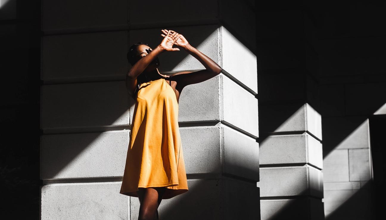 Woman in a yellow dress standing in direct sunlight and covering her eyes from the sun.