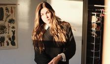 A woman posing for the camera in the sunlight. She is cuffing her shirt.