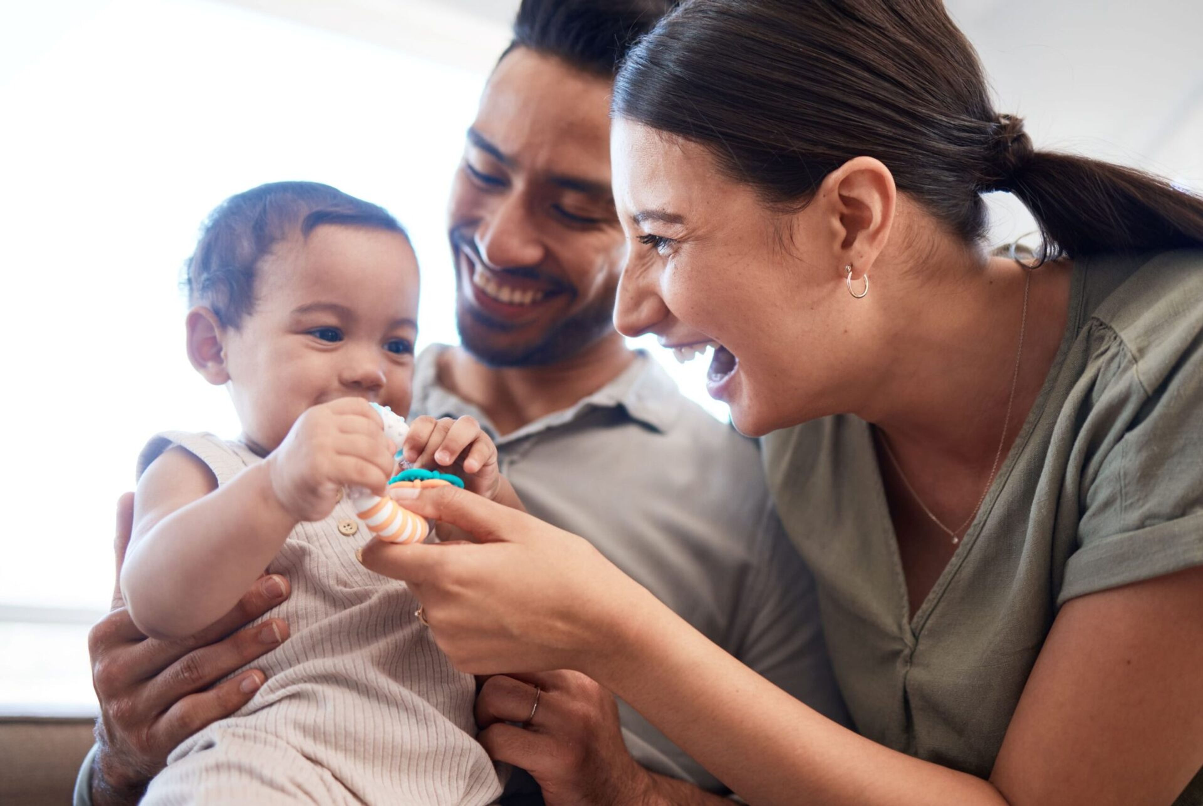 Photo of a father holding an infant while the mother smiles and plays gives the infant a toy