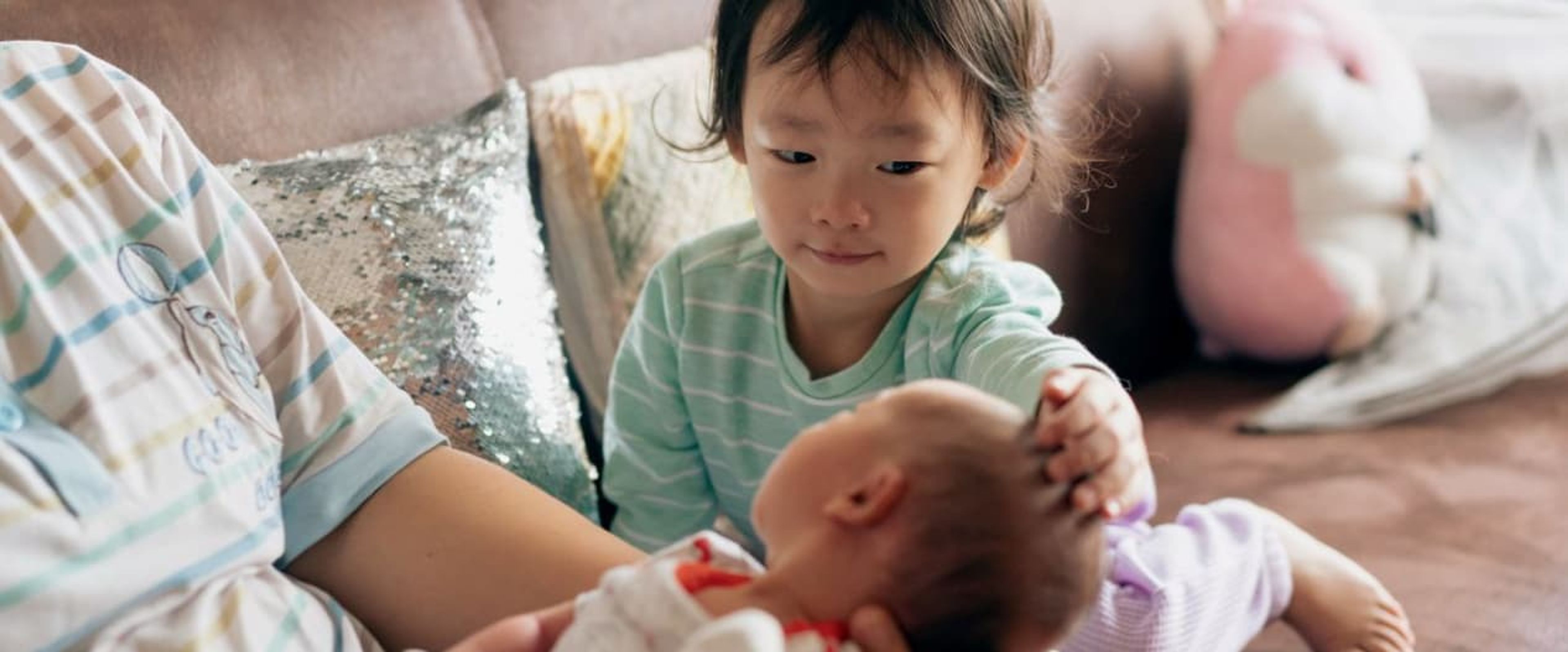Young child gently touching their baby sibling while sitting with their mother.