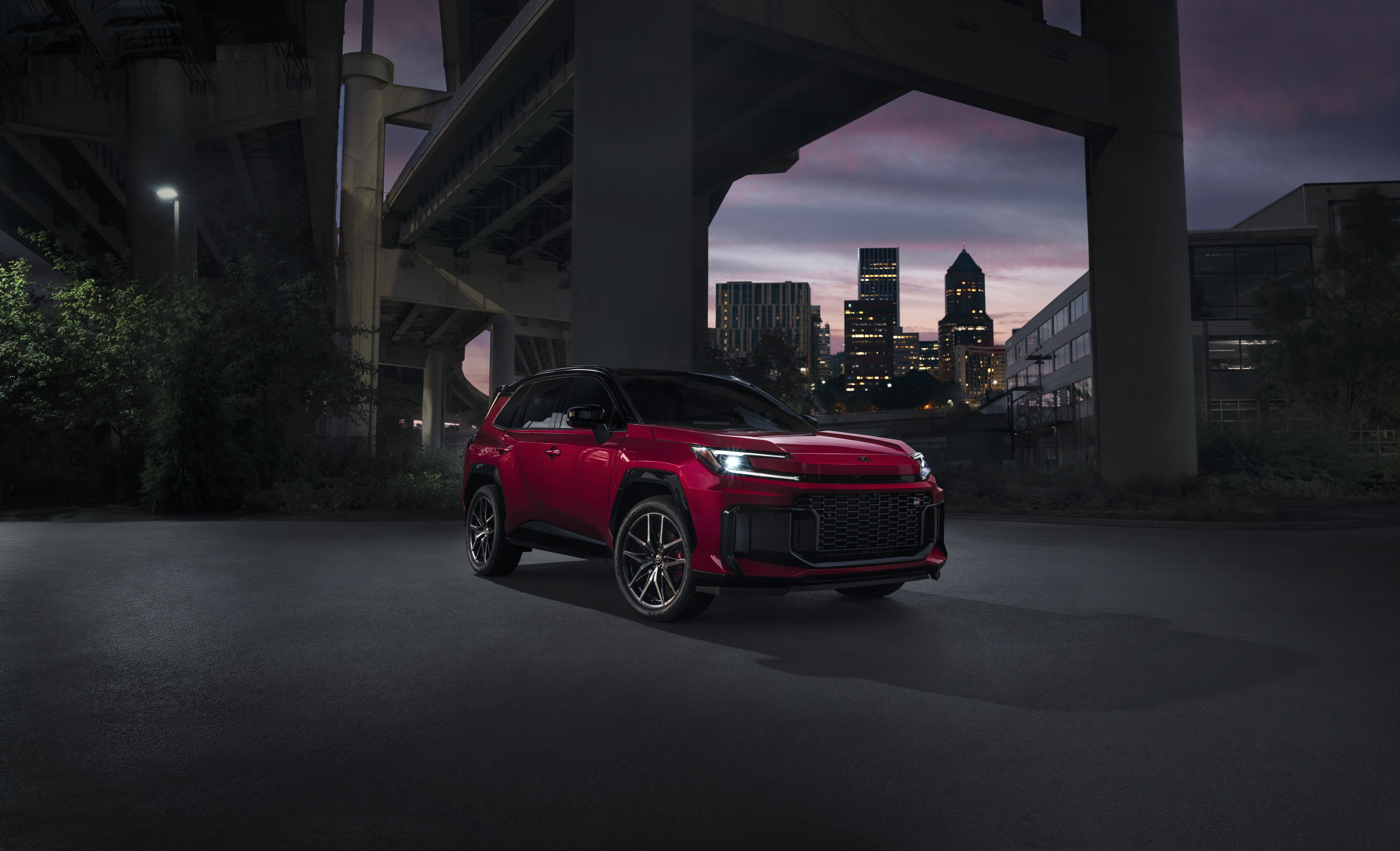 A red Toyota RAV4 parks under a bridge in an urban area with its headlights on