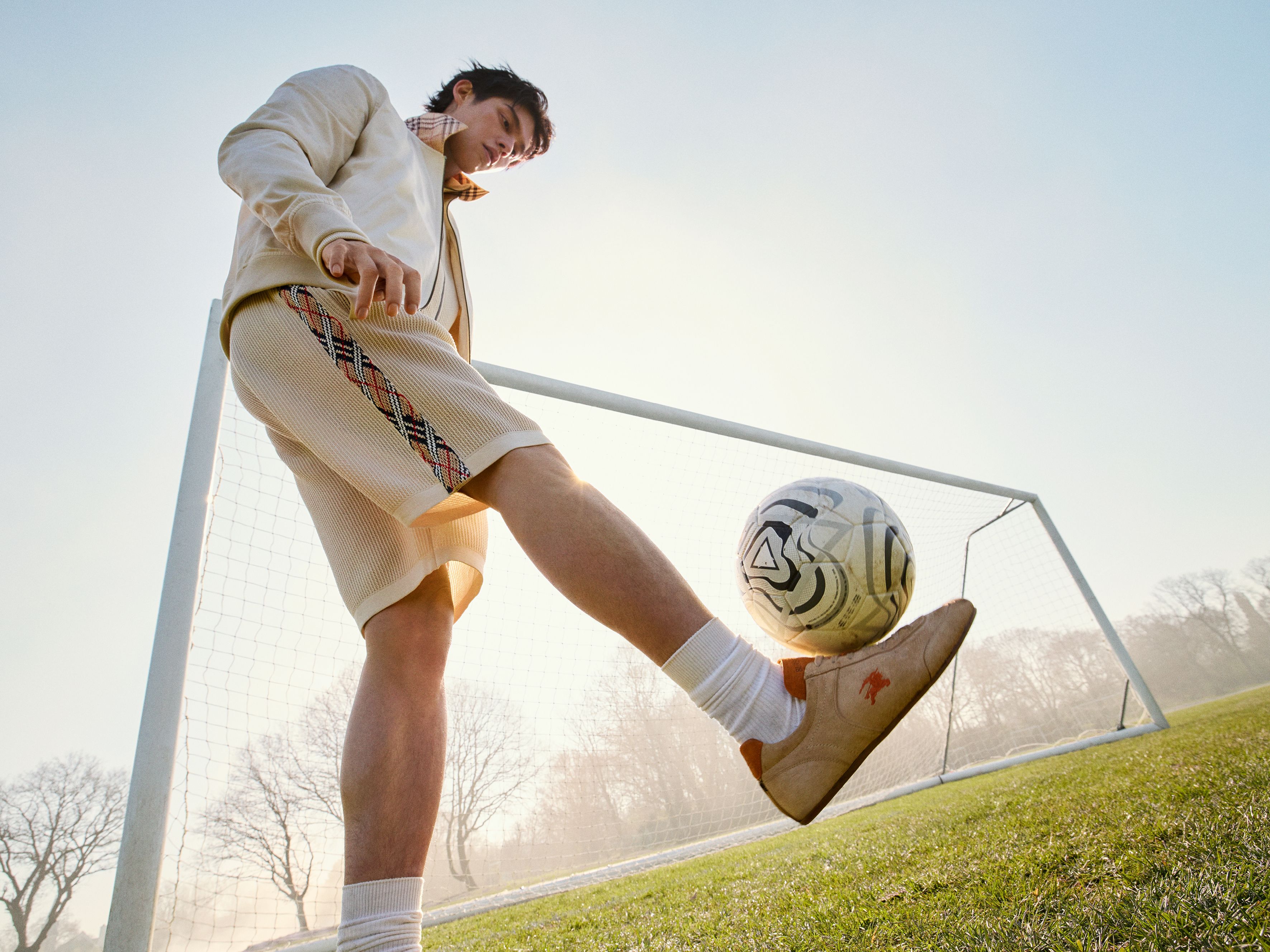 A model dribbles a soccer ball in front of a net in a full Burberry ensemble