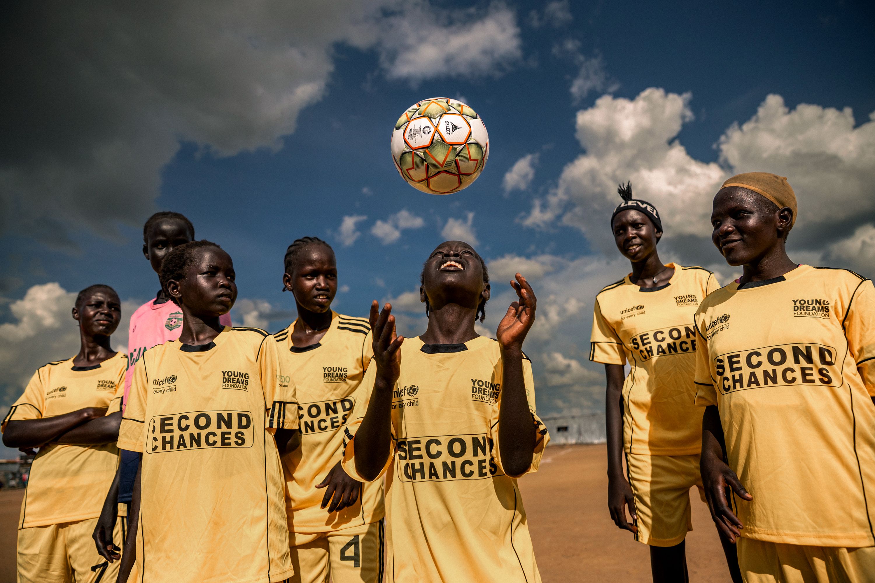 Group of young soccer players in matching yellow jerseys gathered on a dirt field, watching a soccer ball tossed in the air.