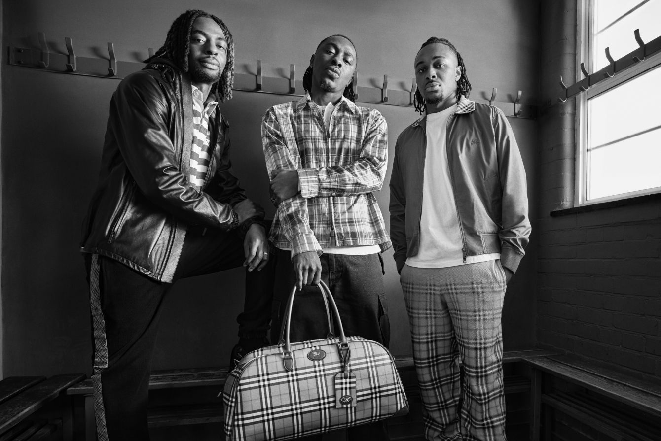 Three people wearing Burberry clothing pose together in a locker room, with one holding a Burberry plaid duffel bag in a black-and-white portrait.
