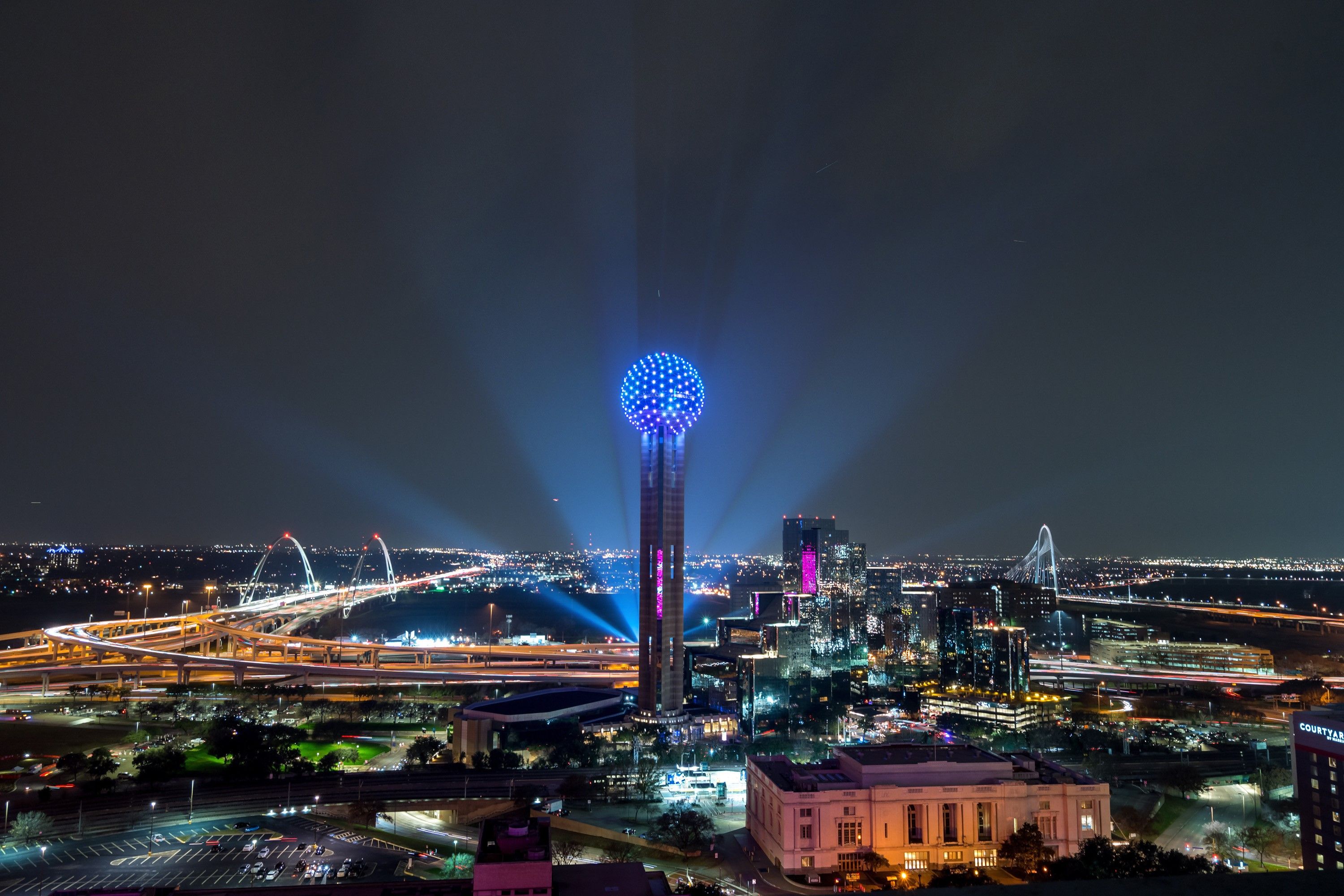 Nighttime view of the Dallas skyline featuring Reunion Tower glowing blue, surrounded by illuminated highways, bridges, and city lights.