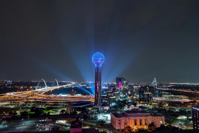 Nighttime view of the Dallas skyline featuring Reunion Tower glowing blue, surrounded by illuminated highways, bridges, and city lights.