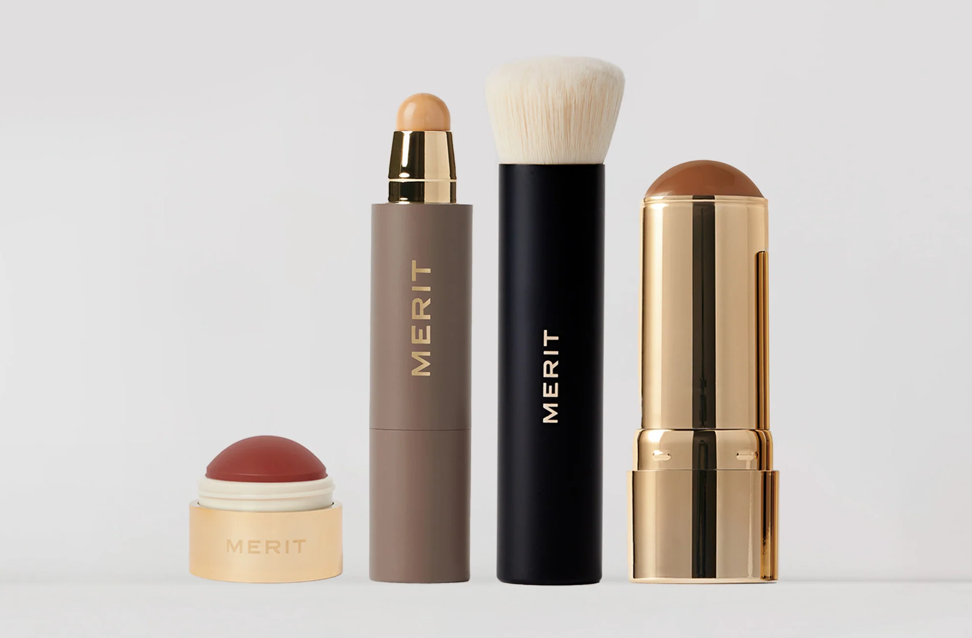 Four MERIT Beauty makeup products displayed against a light background, including a blush balm, complexion stick, brush, and bronzing stick.