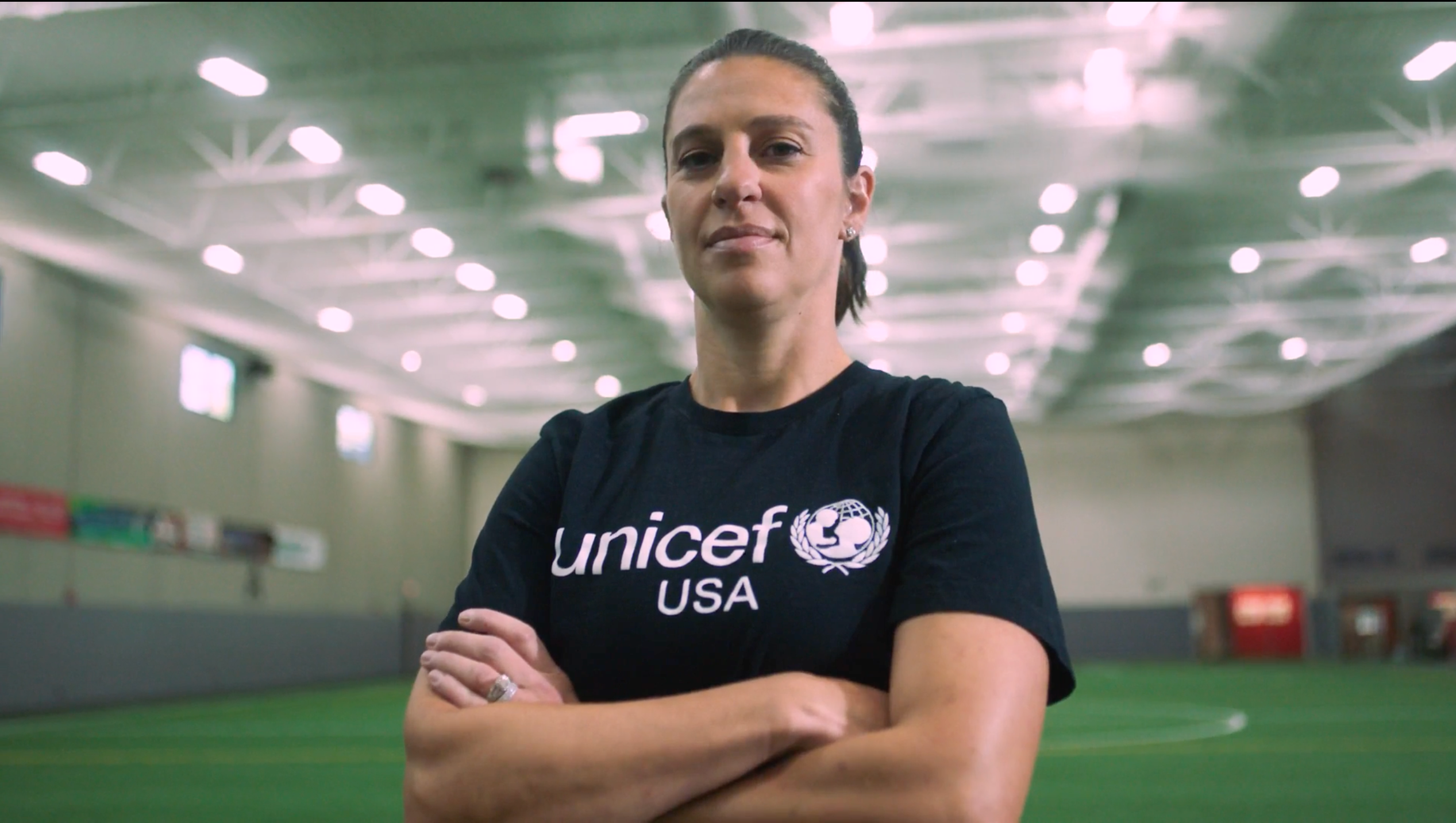 Soccer player Carli Loyd stands with her arms crossed inside an indoor sports facility while wearing a UNICEF USA t-shirt.