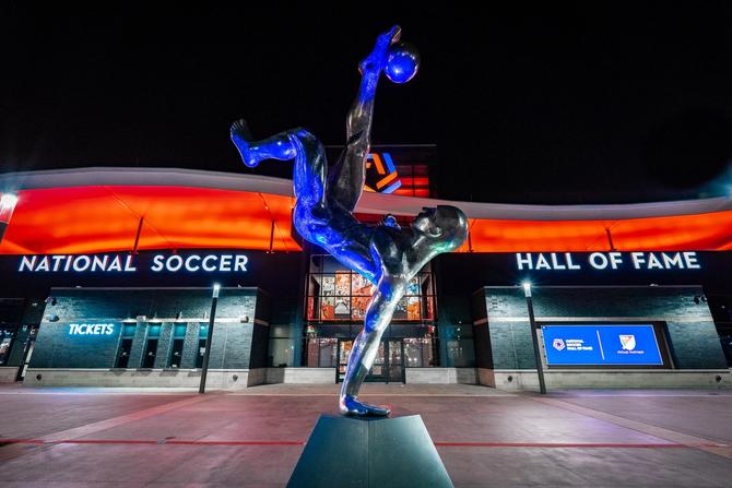 Statue of a soccer player performing a bicycle kick outside the National Soccer Hall of Fame at night.