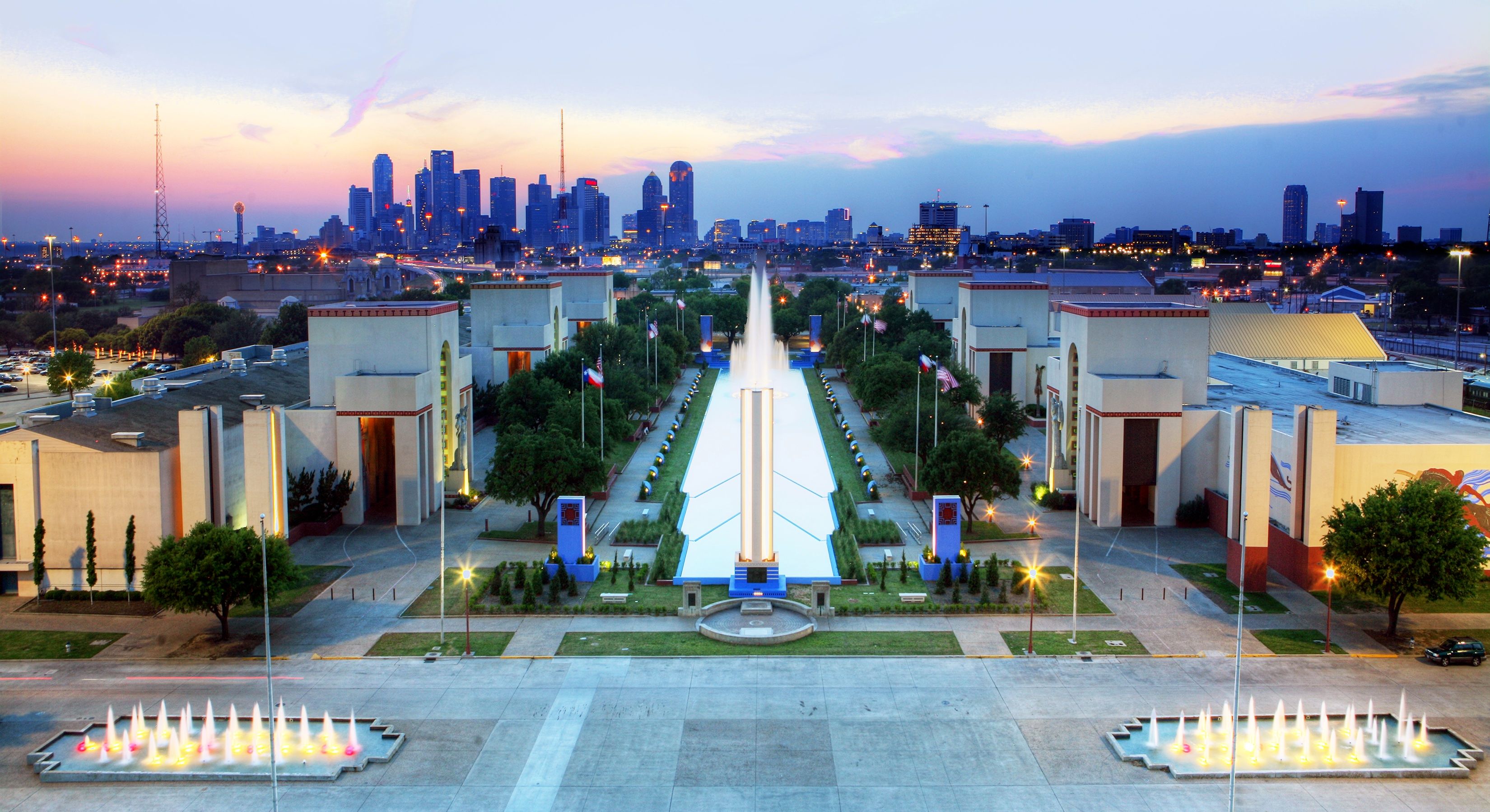Aerial view of Fair Park in Dallas at dusk with the city skyline in the background.
