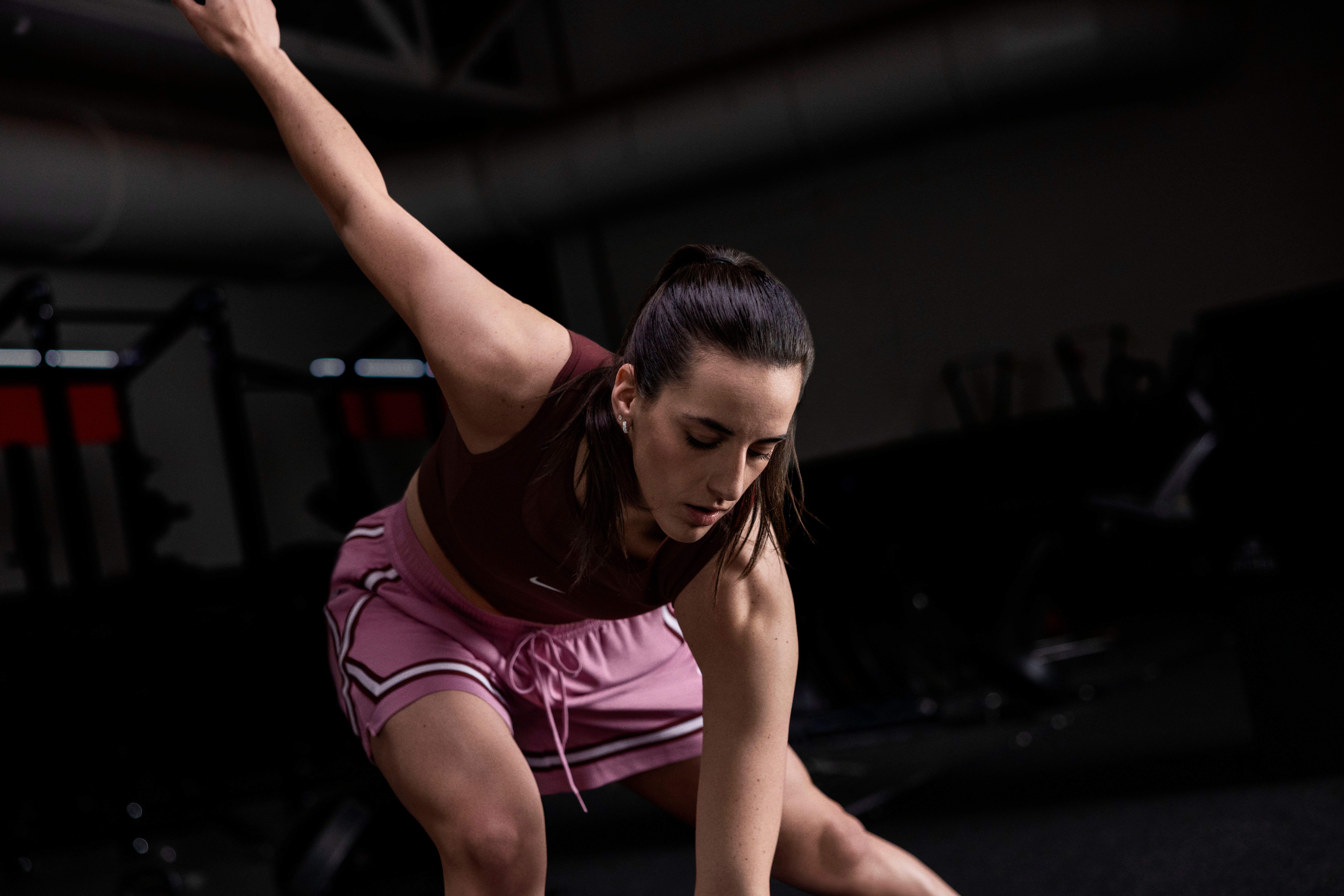 Caitlin Clark stretches in a dark basketball gym wearing pink and red athletic wear