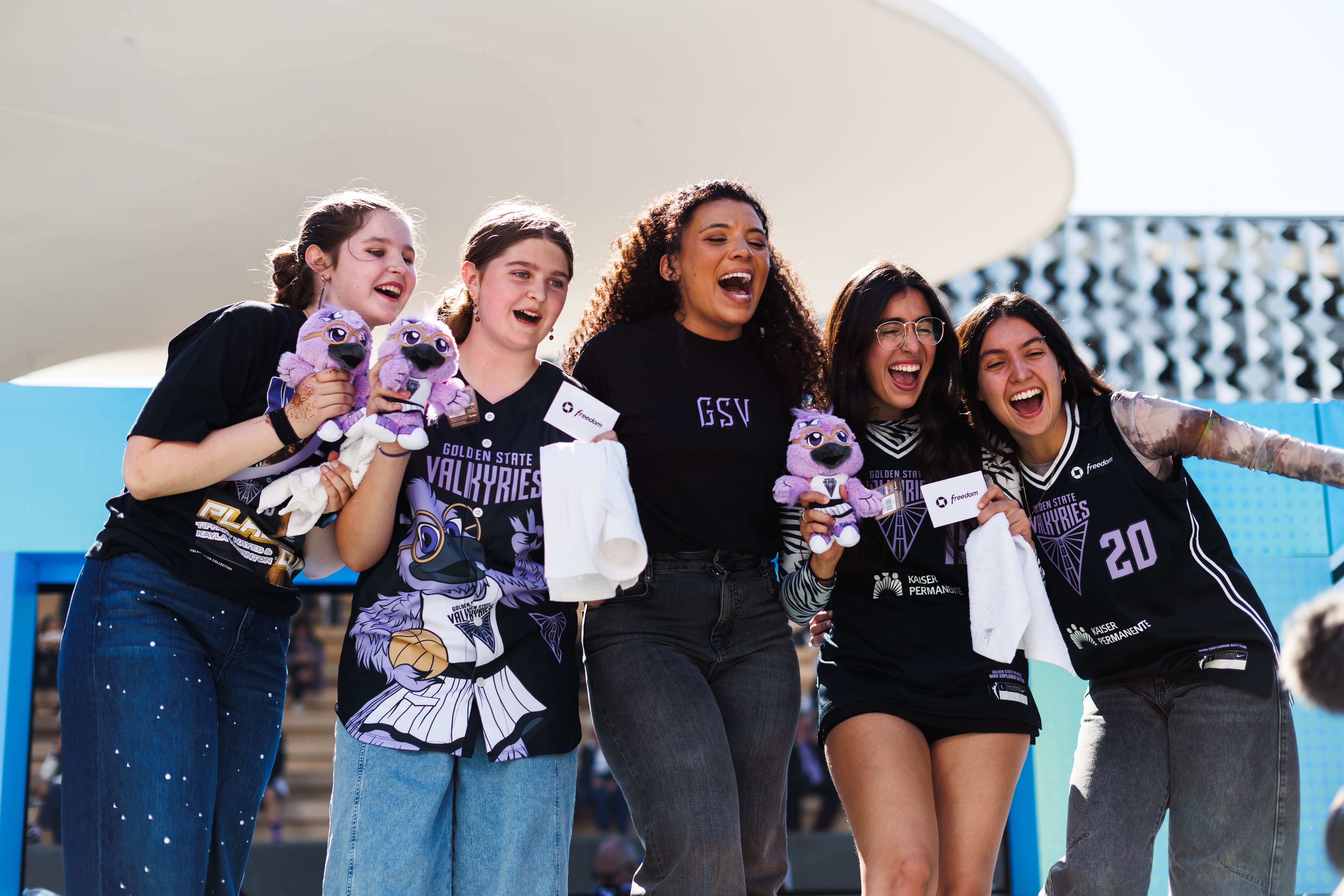 Group of Golden State Valkyries fans smiling and cheering together outdoors, holding team mascot plush toys and rally towels while posing for a photo.