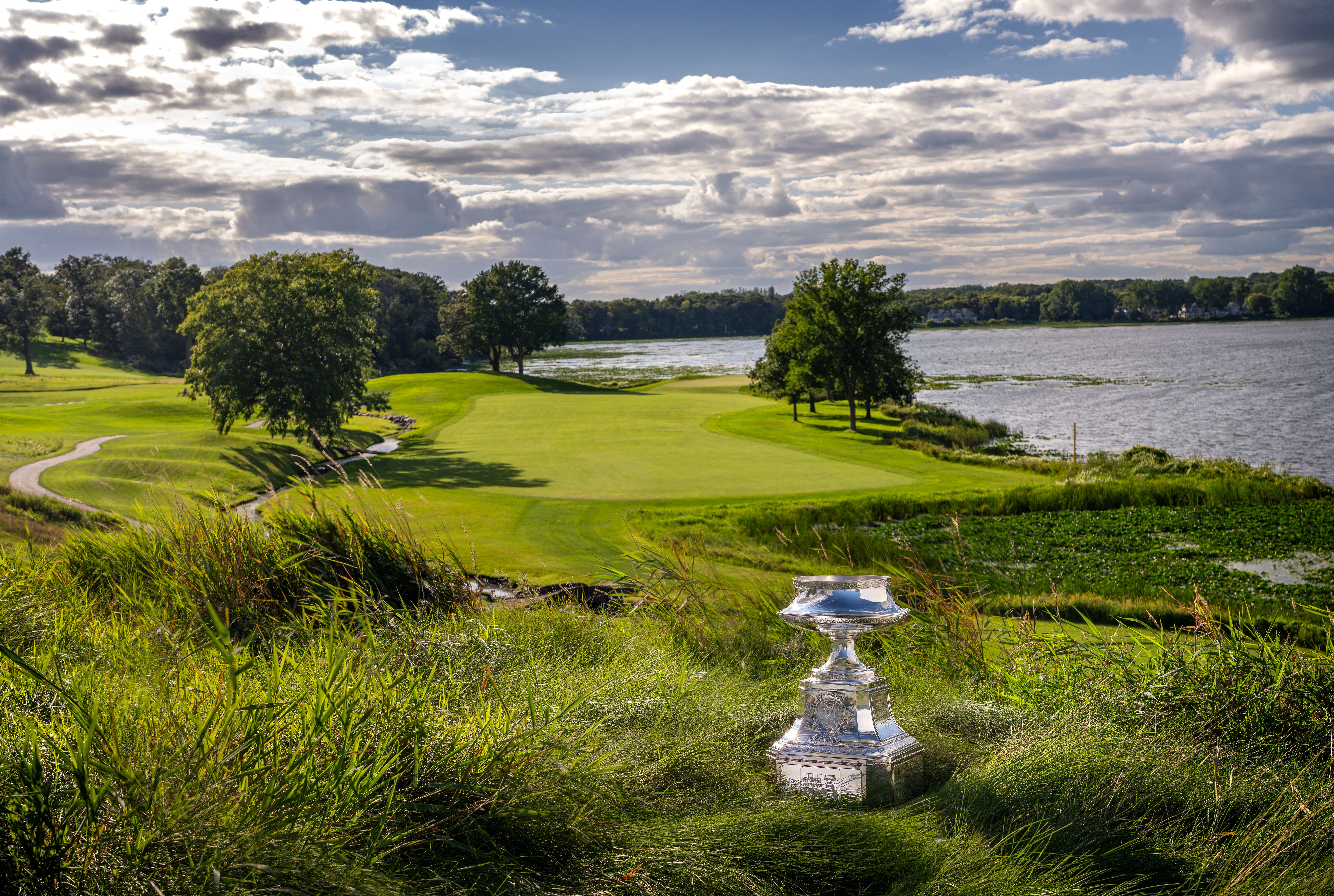 Scenic lakeside golf course under a partly cloudy sky, with the KPMG Women's PGA Championship trophy displayed in the foreground.