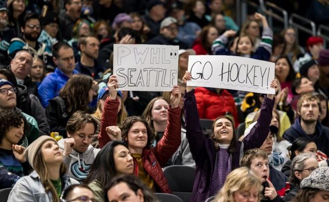 Two fans hold up signs about PWHL Seattle.
