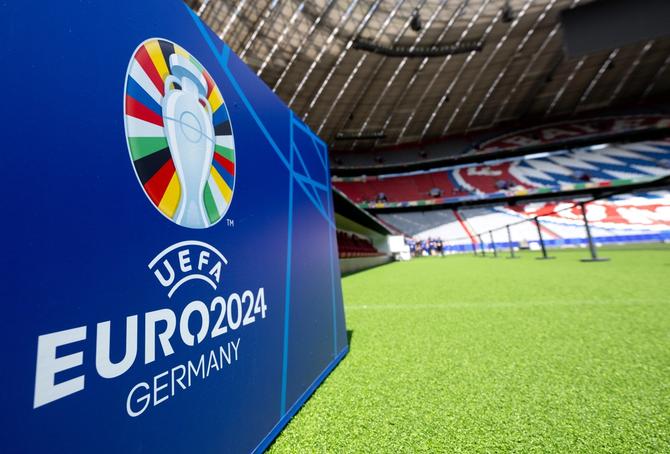 The UEFA EURO2024 logo is seen on a blue wall with Munich Football Arena seating shown in the background.