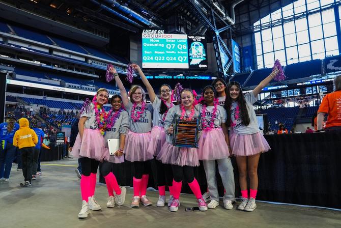 Seven girls dressed in pink stand together for a group photo. Some are holding pink pompoms.