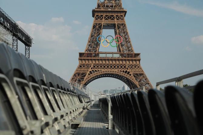 Spectator seats are shown in the forefront of a wide-angle shot of the Eiffel Tower adorned with the iconic six Olympic rings.