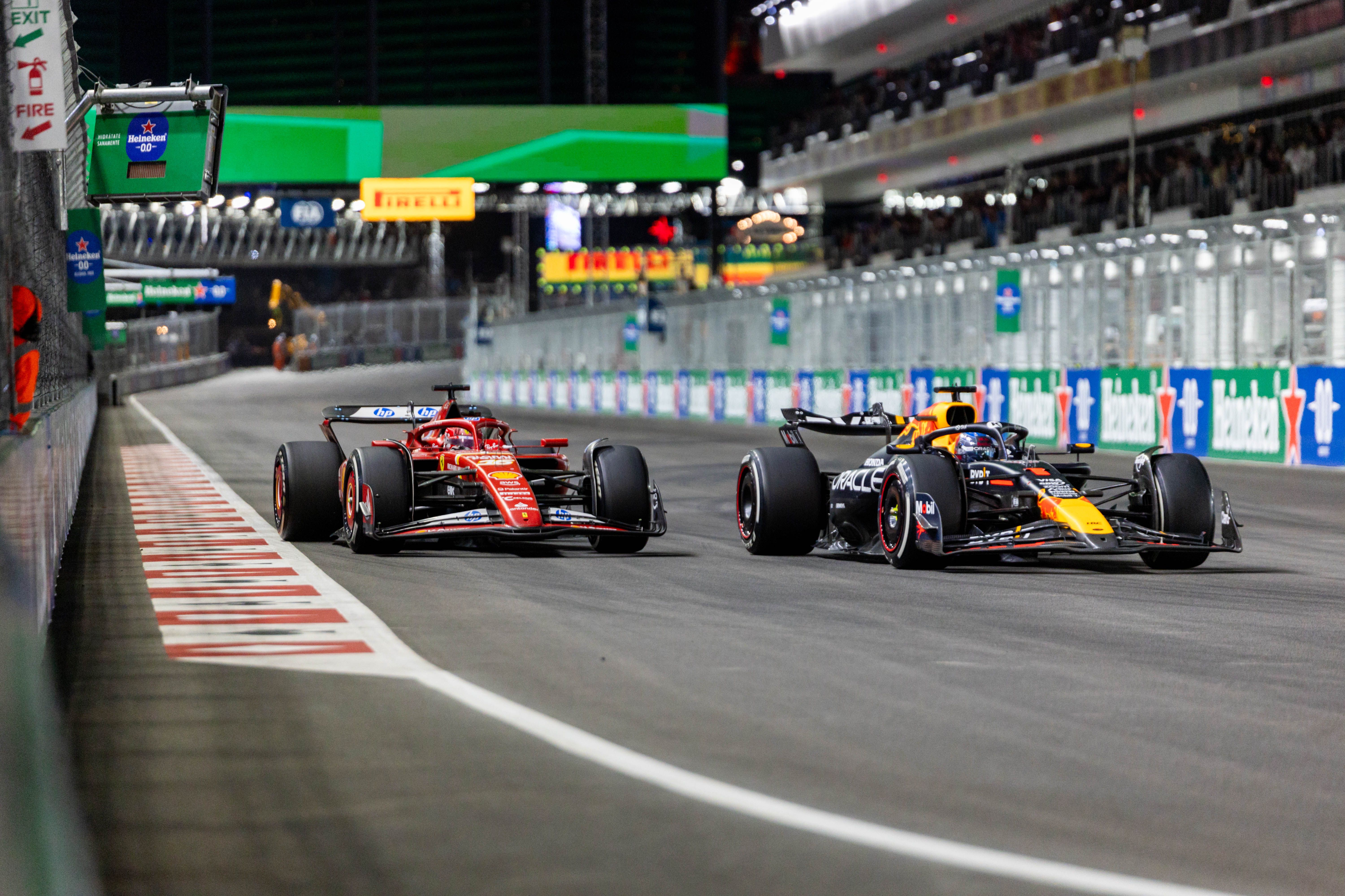 Two Formula 1 cars, a red Ferrari and a dark blue Oracle Red Bull Racing car, racing side by side on a floodlit street circuit at night