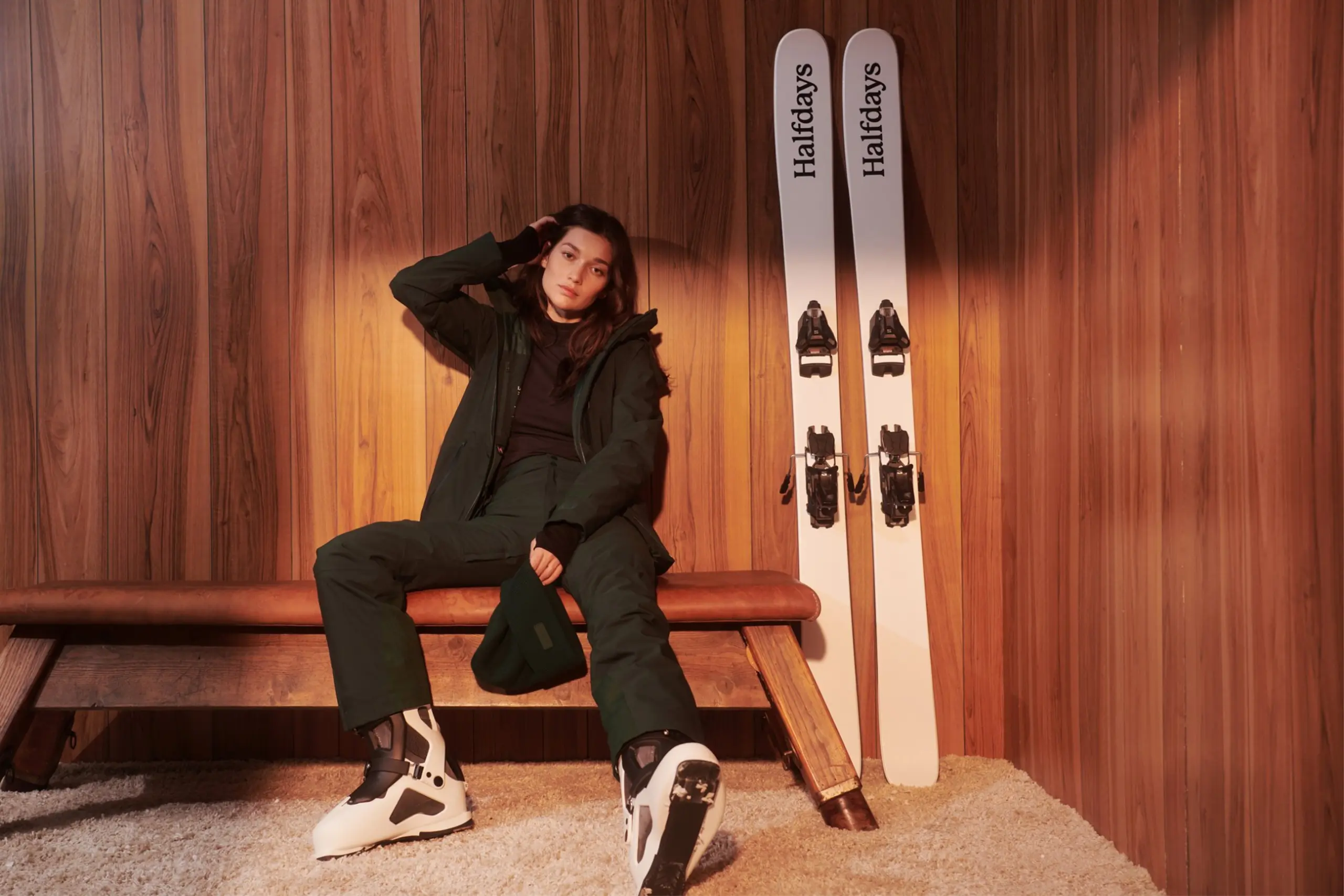 A Halfdays model wears a green ski kit next to a pair of branded skis.