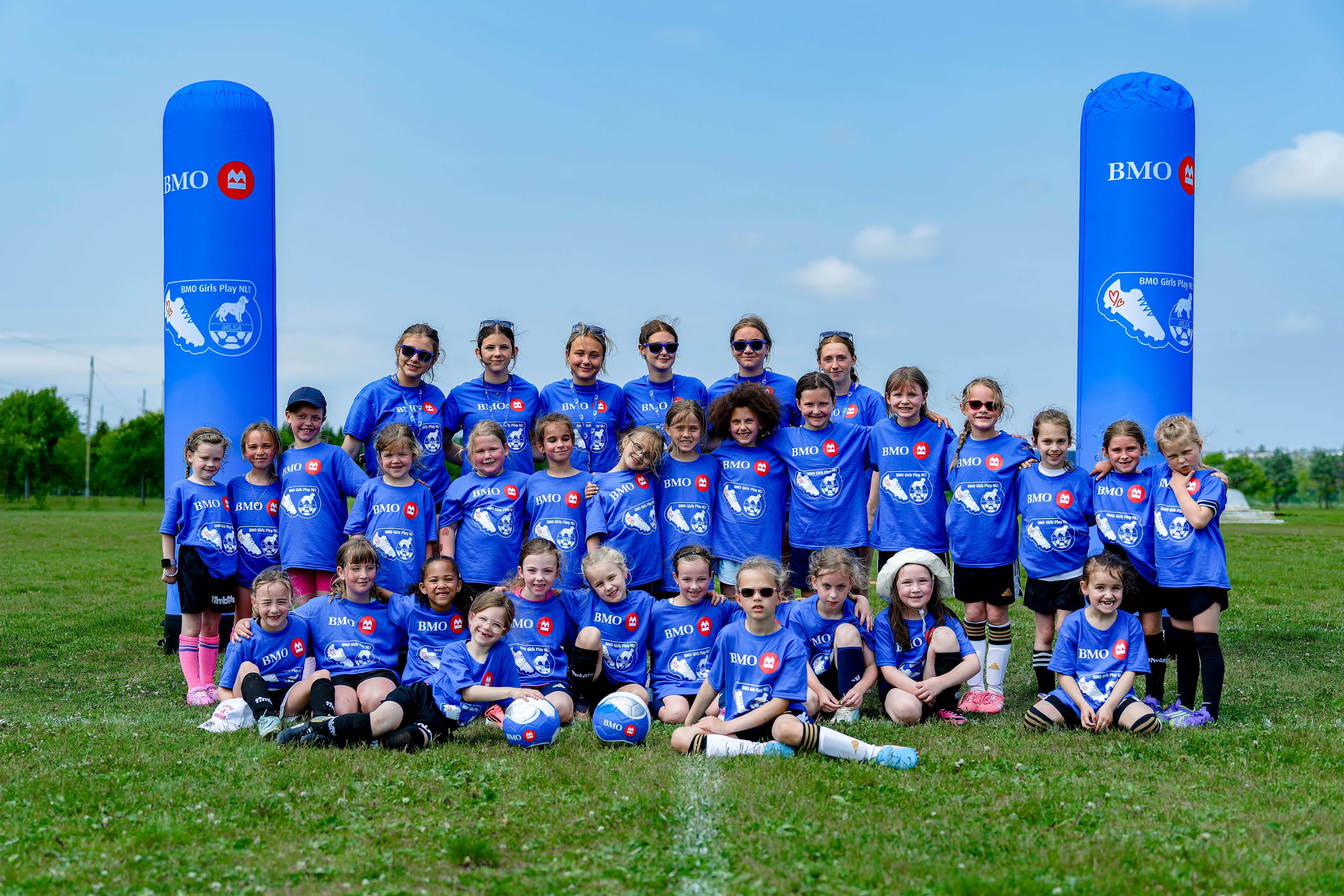 Group photo of youth soccer players and coaches wearing matching blue BMO Girls Play shirts, gathered on a soccer field between branded inflatable pillars during a community soccer event.
