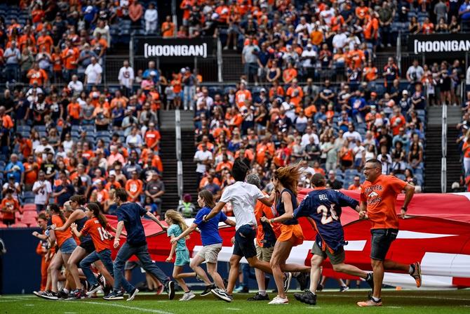 Fans run on the field pulling a field-size American flag at an NFL Football game.