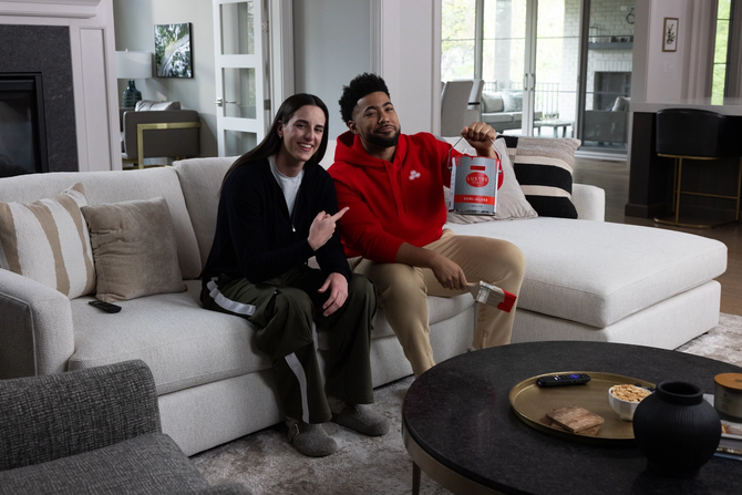Caitlin Clark smiling and pointing at Jake from State Farm while sitting on a couch