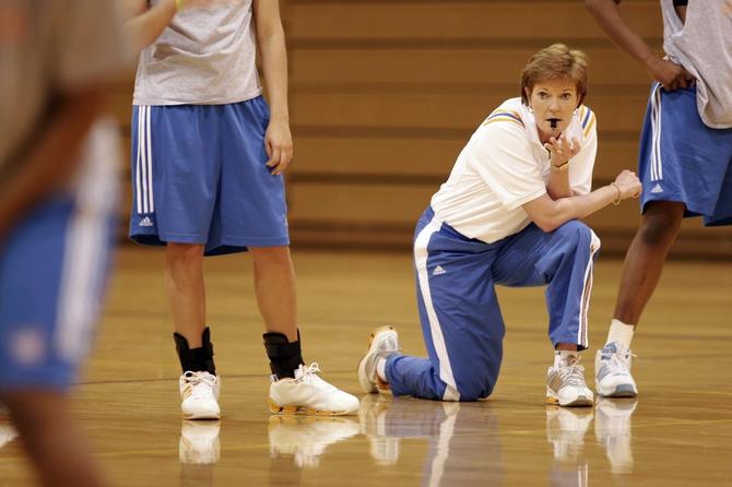 Pat Summitt kneels on the ground during practice with a whistle in her mouth, looking on.