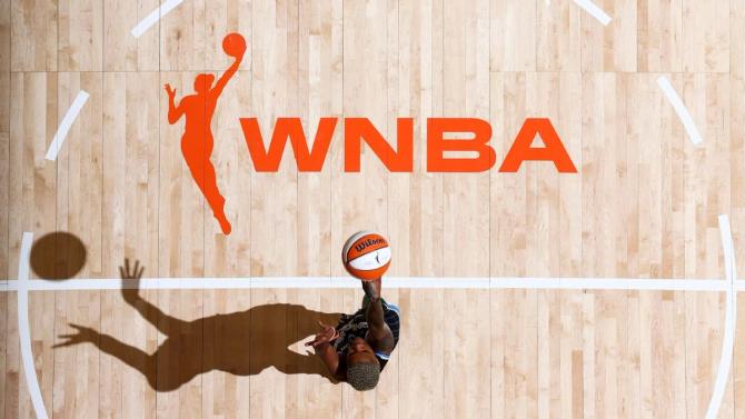 An overhead shot of the WNBA logo on the court with a woman shooting a basketball.