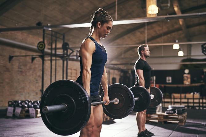 A woman stands in the foreground whie a man stands in the background. Both athletes are holding a barbell and preparing to complete a lift.