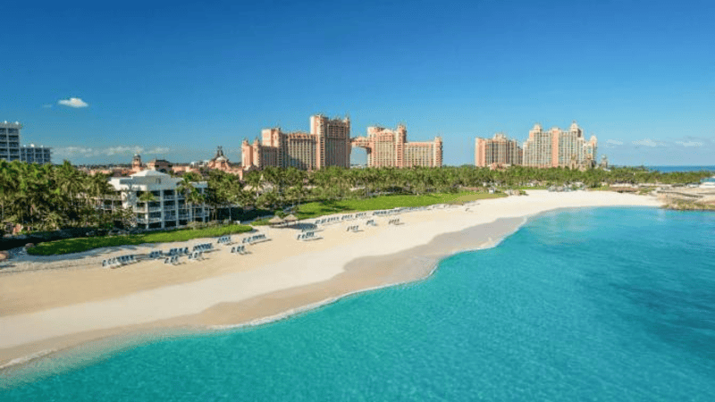 Aerial view of Atlantis resort with large pink hotel buildings, palm trees, white sand, rows of lounge chairs and clear turquoise ocean water under a bright blue sky.