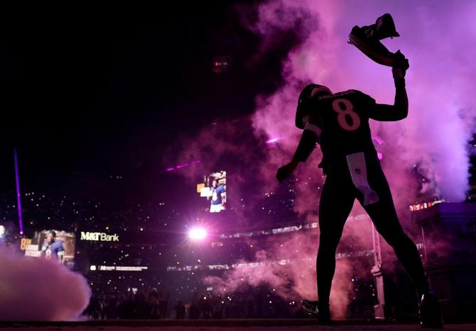 Alt text: Lamar Jackson walks into his home stadium waving a towel as smoke surrounds him.