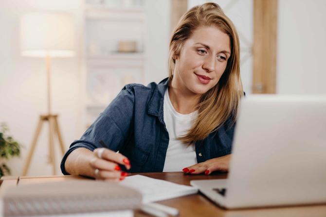 A woman staring at her laptop while taking notes on a notepad at her desk